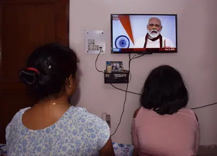 Eine Frau und ein Mädchen sitzen vor einem TV, in welchem Narendra Modi zu sehen ist. 